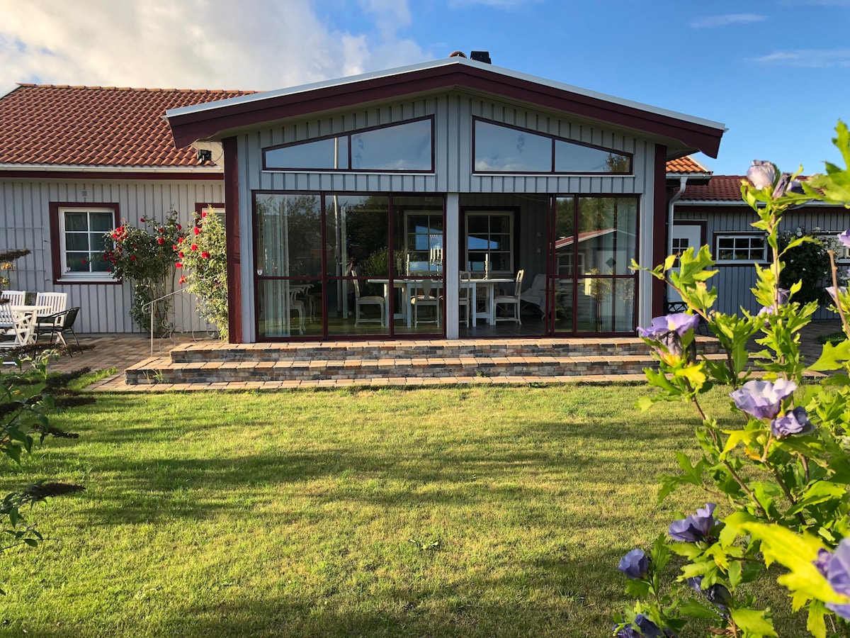A charming house features a spacious, sunlit terrace with large glass doors leading into a welcoming interior. The garden is adorned with blooming flowers, while a neatly paved area invites outdoor gatherings. A peaceful green lawn extends around the property.