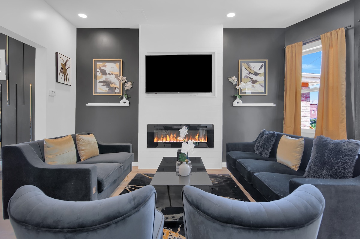 A modern living room features two plush, dark couches and two accent chairs arranged around a sleek coffee table. A wall-mounted flatscreen TV is above an elegant fireplace, with artwork displayed on adjacent walls. Large windows with yellow curtains provide natural light.