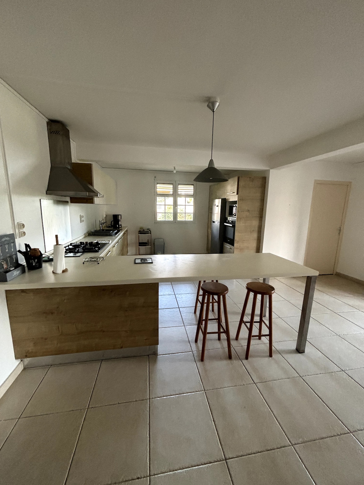 A spacious kitchen features a long countertop with two wooden stools positioned nearby. Stainless steel appliances and a range hood are installed above the cooking area. Natural light comes through a window, illuminating the tiled floor and surrounding cabinetry.