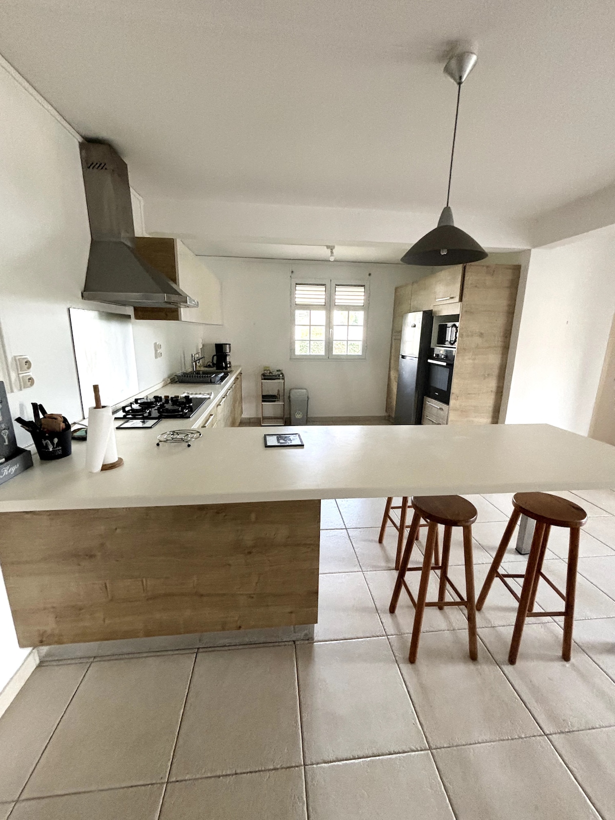 A spacious kitchen is equipped with modern appliances, including a gas stove and refrigerator. A central island with a smooth countertop is accompanied by two wooden stools, providing a casual dining space. Natural light enters through the window, illuminating the neutral-toned cabinetry and tiled floor.