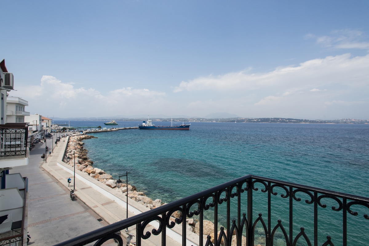 A scenic view of the harbor and sea is presented from the furnished balcony. Clear blue waters reflect the sky, while a ship is anchored nearby. The shoreline is lined with rocky formations and a paved walkway, adding to the coastal charm.