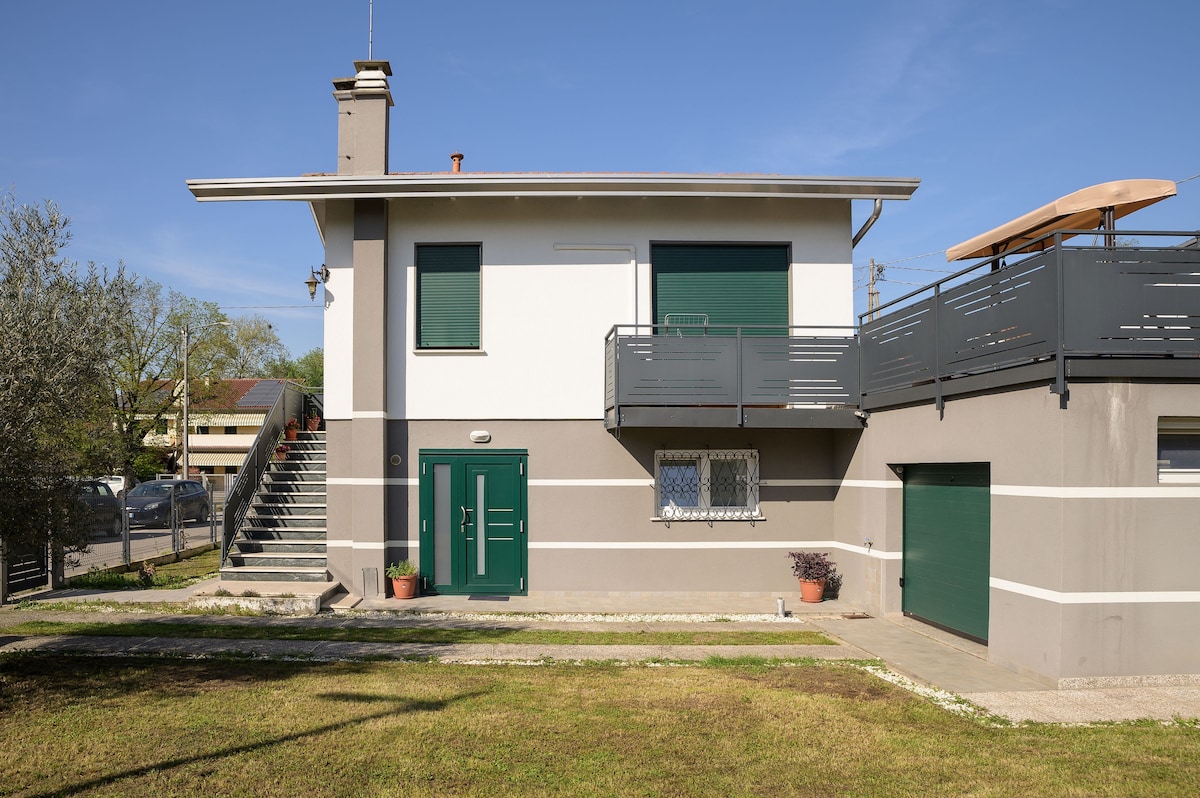 The exterior of a two-story villa is showcased, featuring a green door framed by a concrete facade. A staircase leads to the upper level with a balcony and a sun lounger. A driveway to the garage is visible, along with landscaped grass.