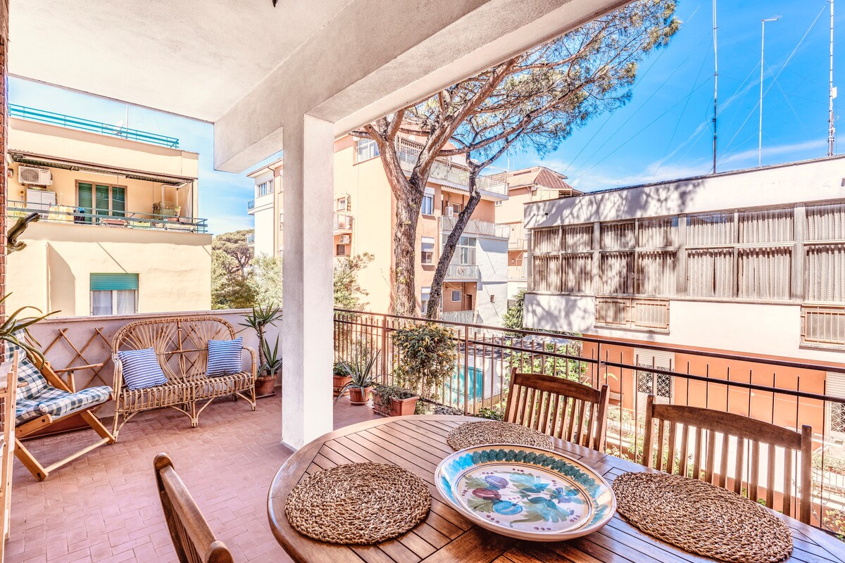 An outdoor terrace is featured, showcasing a wooden dining table surrounded by woven chairs. Lush greenery and potted plants are visible, enhancing the inviting space. The scene is complemented by a clear blue sky, with apartment buildings in the background.