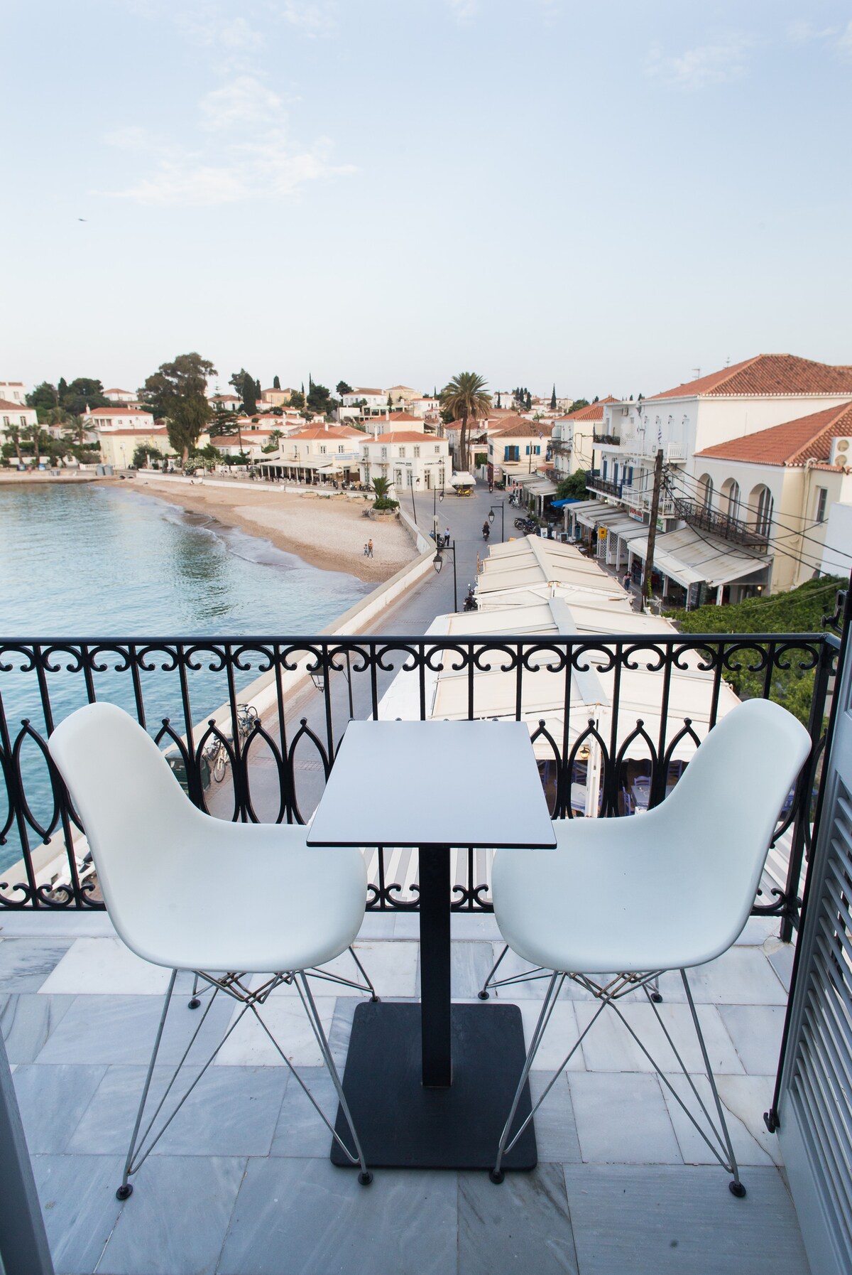 A small balcony features a round table with two white chairs, providing a view of a sandy beach and waterfront lined with palm trees. The picturesque village scene includes charming buildings and outdoor dining spaces, enhancing the serene coastal atmosphere.
