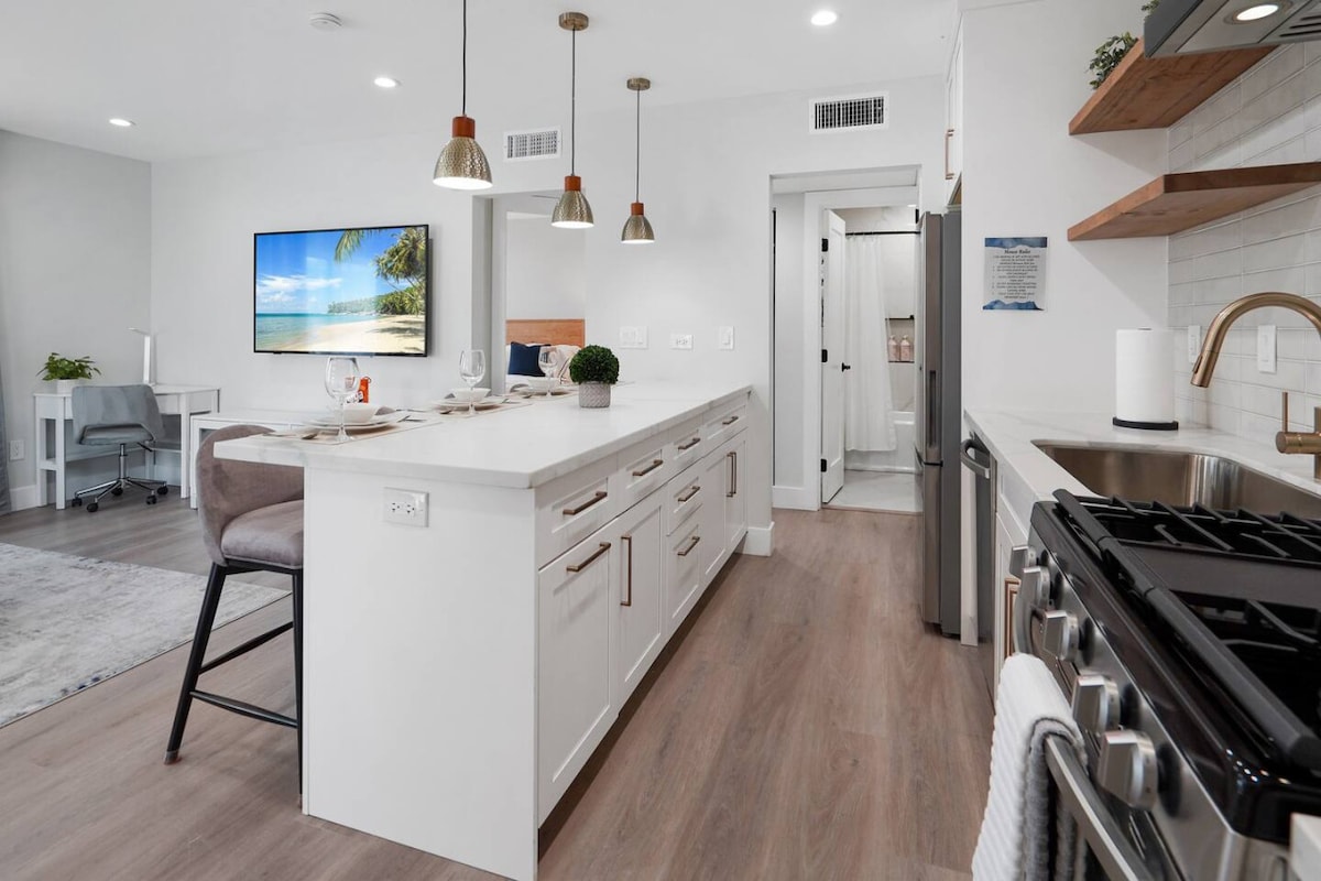 A modern kitchen features sleek white cabinetry and a spacious island with bar stools. A 65-inch smart TV is mounted on the wall, providing entertainment. Natural light enhances the inviting atmosphere, while stainless steel appliances are visible alongside stylish open shelving.