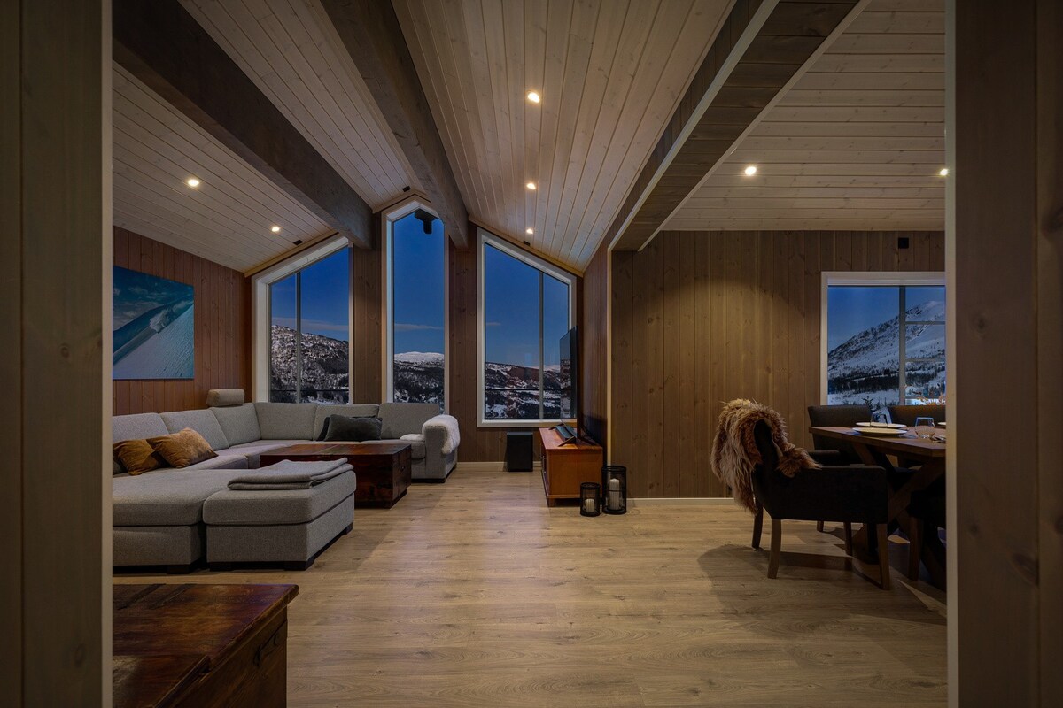 A modern living area features high, sloped ceilings and large panoramic windows offering views of the surrounding landscape. A comfortable sectional sofa and wooden coffee table occupy the space, while a dining table is positioned nearby, allowing for shared meals.