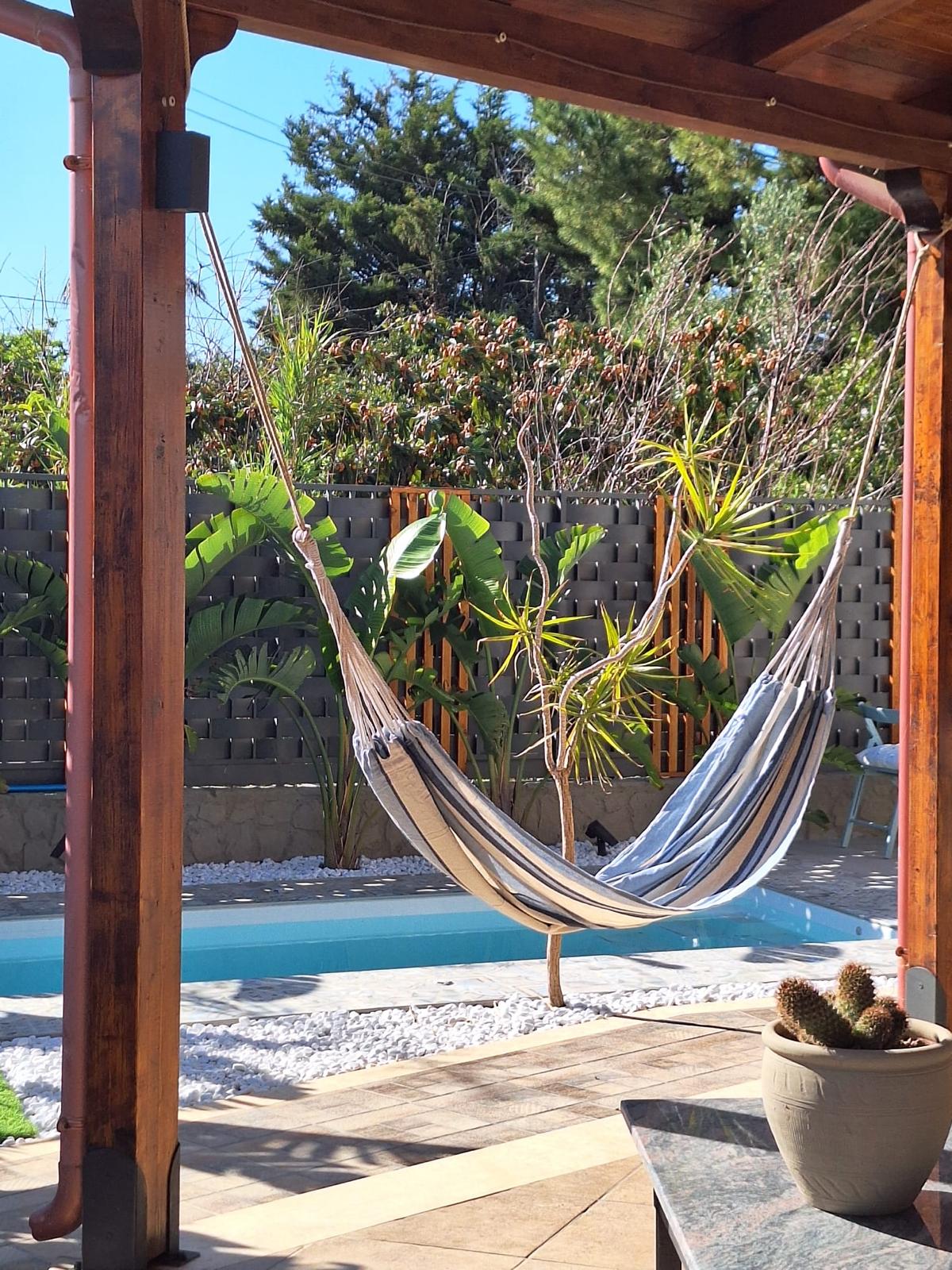 A serene outdoor space is showcased, featuring a striped hammock suspended under a wooden pergola. In the background, a small pool is surrounded by decorative stones and lush greenery, providing a tranquil retreat for relaxation.