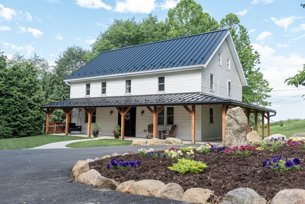 The newly renovated farmhouse, featuring a gabled roof and a wrap-around porch, is surrounded by lush greenery and colorful flower beds. The entrance is framed by large stones and a well-maintained driveway that leads directly to the porch area.