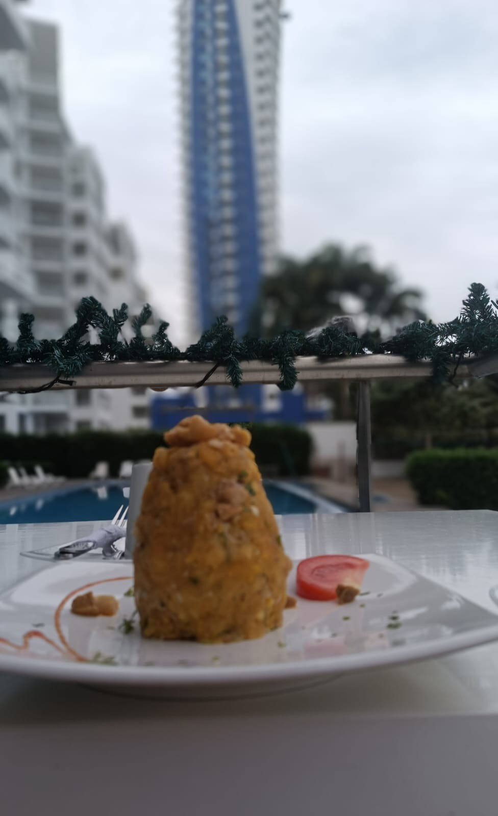 A neatly presented dish is displayed on a white plate, featuring a tall mound of food decorated with herbs and garnishes. In the background, a pool area is visible, along with modern buildings and greenery, creating a relaxed dining atmosphere.