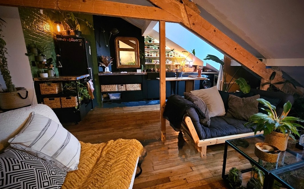 The image showcases a cozy loft space with a wooden frame and sloped ceiling. A seating area is highlighted by plush cushions and a striped throw. A mirrored vanity and a kitchen area with plants and shelves are visible in the background, creating an inviting atmosphere.