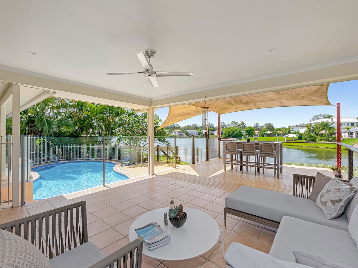 A spacious covered patio provides seating areas with modern outdoor furniture, including a sectional sofa and coffee table. A private swimming pool is visible in the foreground, while the serene lake and lush greenery serve as a backdrop, creating a relaxing outdoor environment.