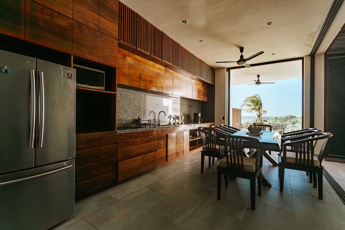 A modern kitchen features wooden cabinetry and a sleek countertop, paired with stainless steel appliances. A dining area with a large table and multiple chairs is positioned near expansive sliding doors that provide a view of the outdoors and allow natural light to fill the space.