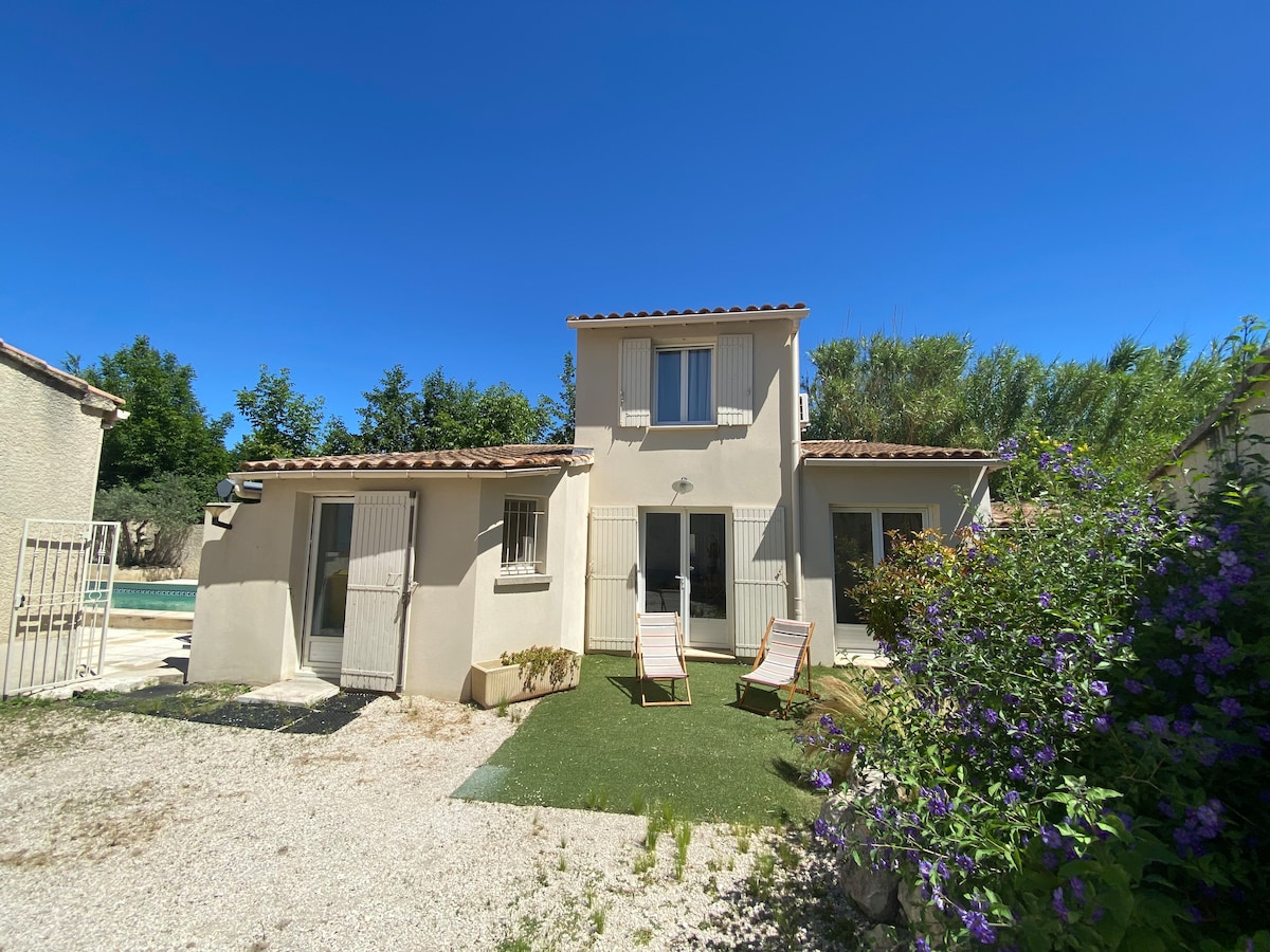 A charming vacation home is set against a clear blue sky, featuring a light-colored façade with shutters. Two lounge chairs are positioned on a grassy area in front, surrounded by blooming plants. A glimpse of the shared pool is visible to the left.