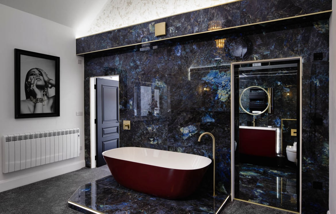 A modern bathroom is featured with a freestanding bathtub in deep red, set against dark marbled walls. A large mirror with a circular frame reflects the space. Stylish lighting enhances the luxurious feel, with a door leading to another area visible at the back.
