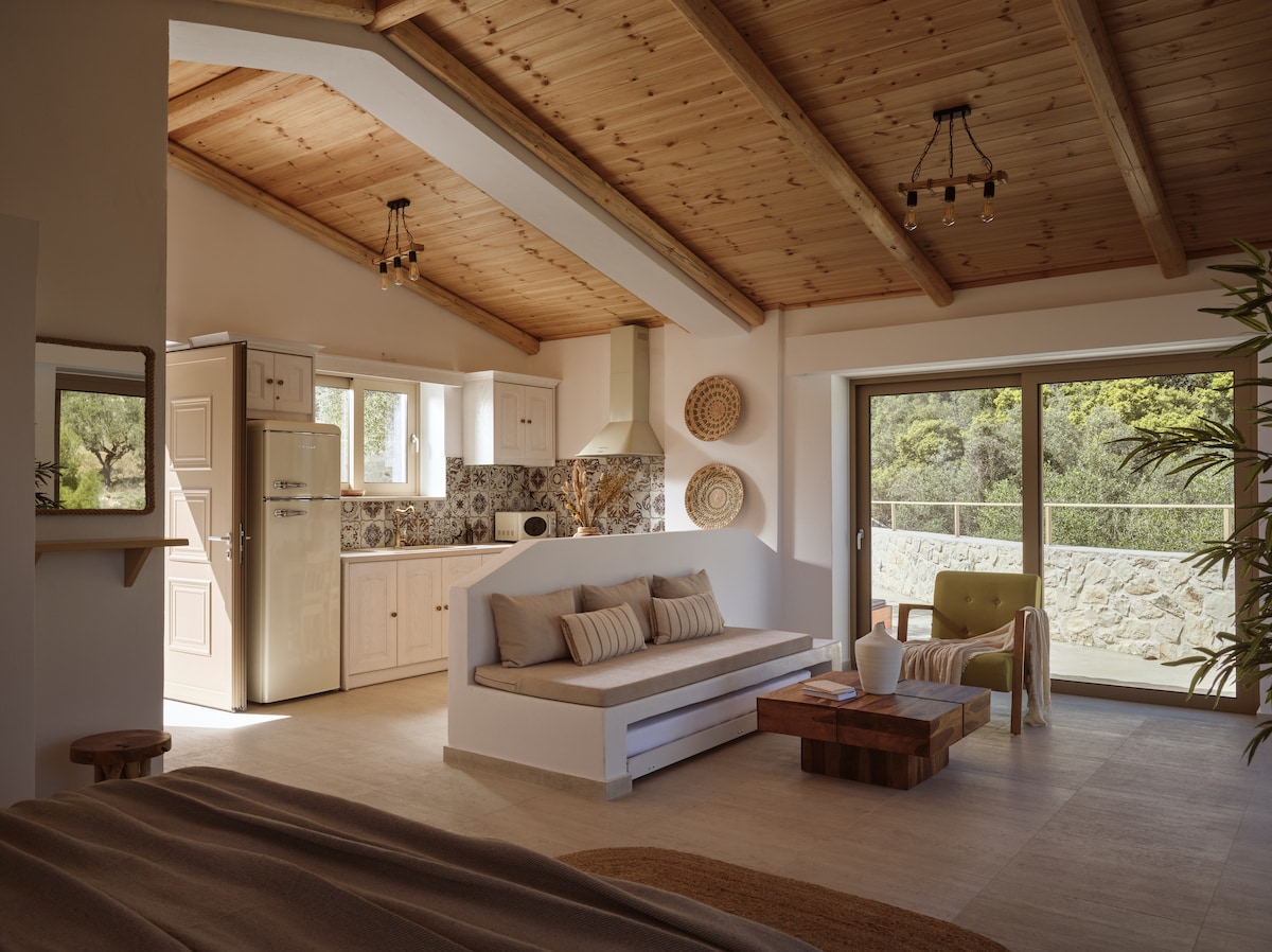 An open living area features a wooden ceiling with exposed beams. A cozy seating arrangement includes a light sofa and a green chair, complemented by a wooden coffee table. Natural light pours in through large glass doors leading to a balcony with scenic views.