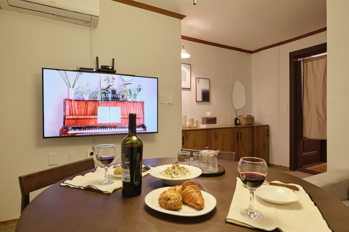 A dining space featuring a round table set with plates and glasses, displaying a bottle of wine and various pastries. A large screen mounted on the wall showcases a digital image of a piano. Soft ambient lighting is provided by a ceiling fixture.