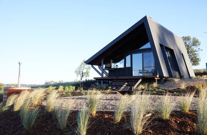  The image shows a modern, angular cabin perched on a hillside with large glass windows offering stunning views. Surrounded by grasses and native plants, it has a sleek, minimalist design that blends perfectly with the natural environment