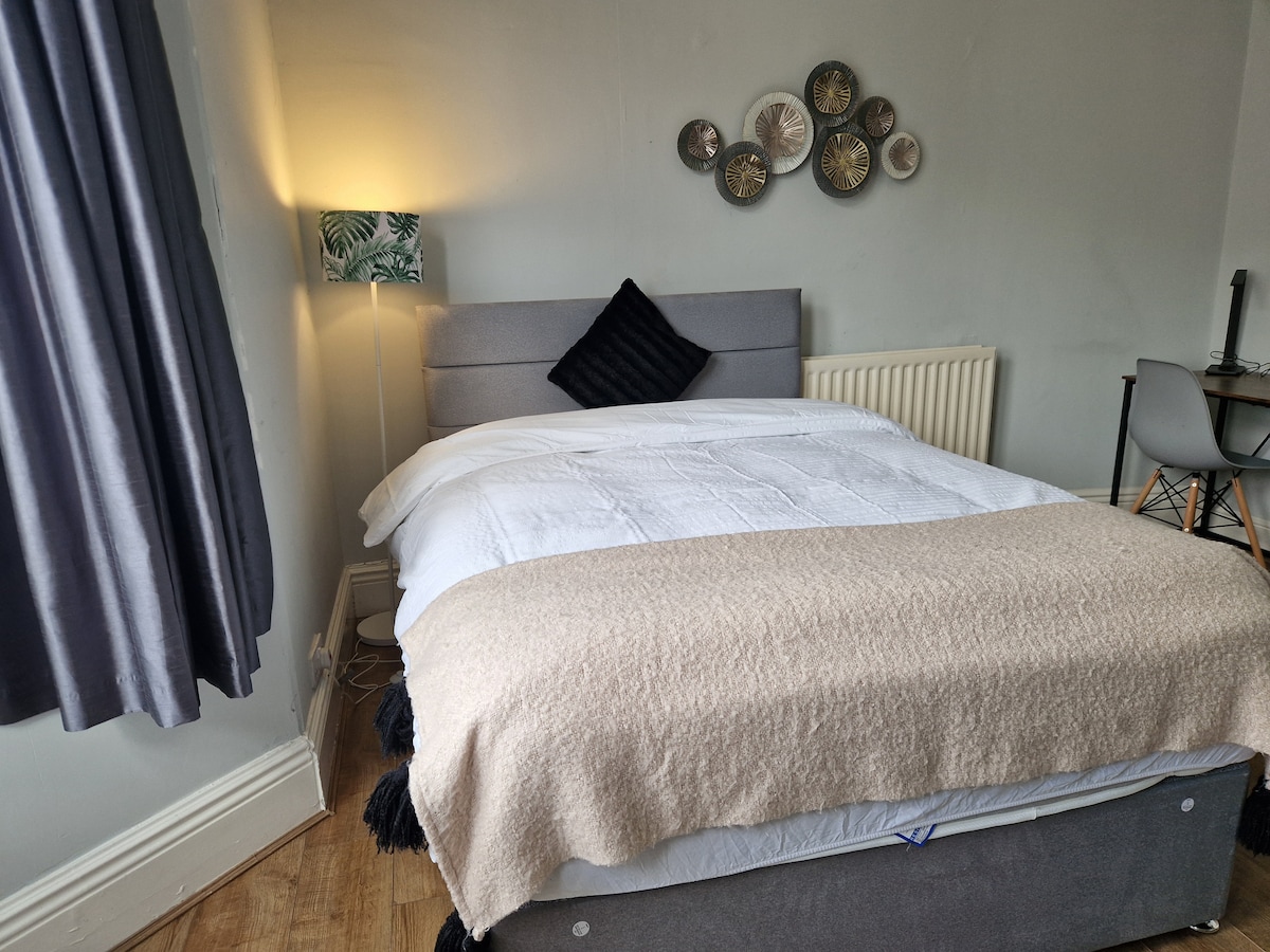 A cozy double room features a spacious bed made with fresh linens, a decorative throw blanket draped across it, and soft lighting from a bedside lamp. A wall-mounted arrangement of decorative plates adds visual interest, while a small workspace with a chair is positioned nearby.