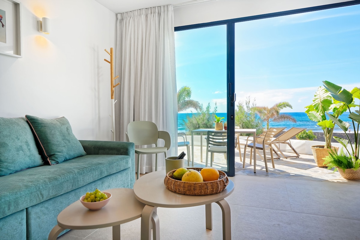 A living area features a soft, turquoise sofa facing large glass doors that open to a terrace with views of the beach. Two coffee tables, one round and one oval, are placed in front of the sofa. Natural light floods the space, highlighting indoor plants.