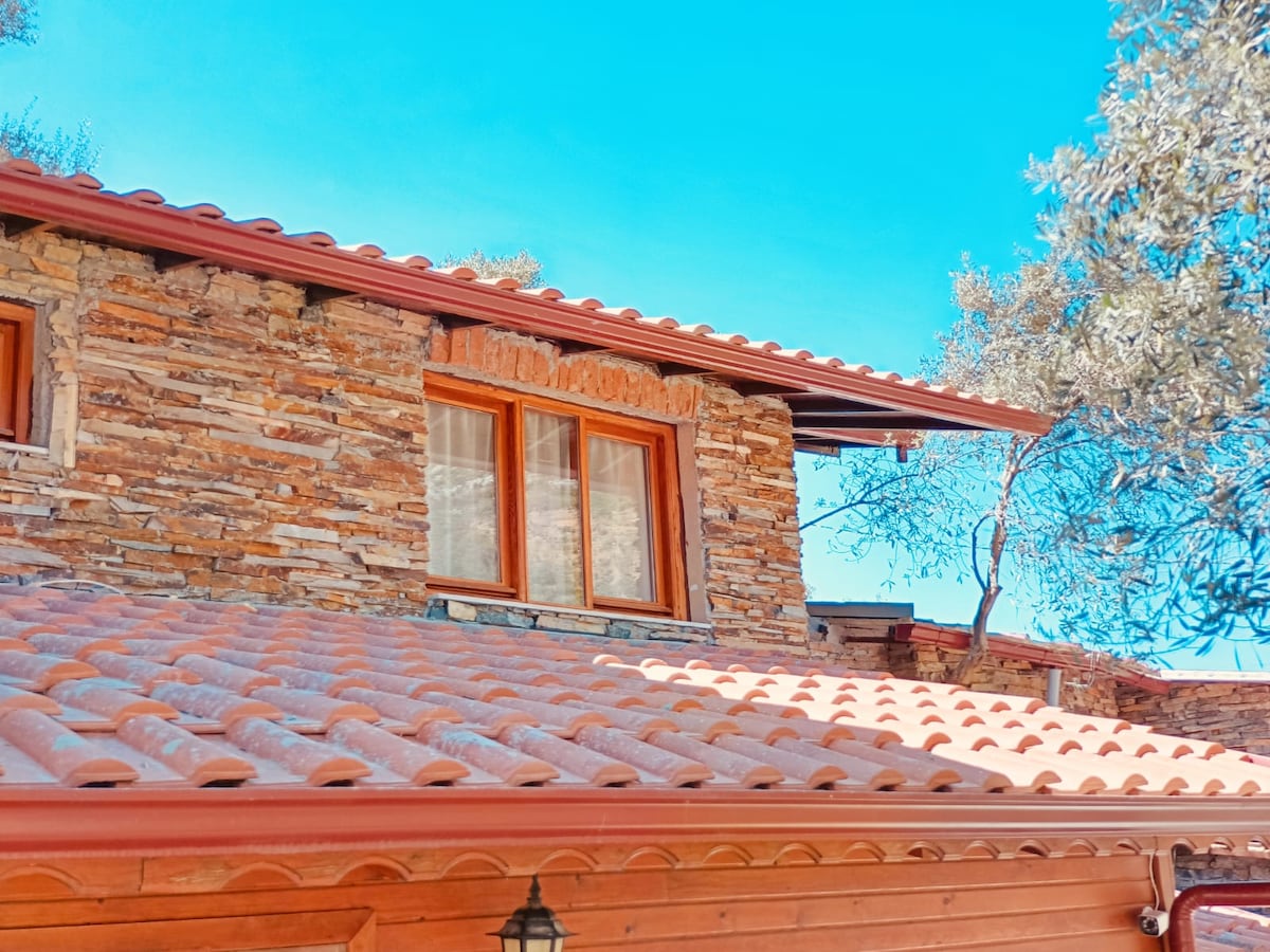 The image features a rustic stone building topped with terracotta tiles under a clear blue sky. Large windows with wooden frames are visible, reflecting the sunlight. Nearby, green foliage adds natural elements to the structure's warm tones.