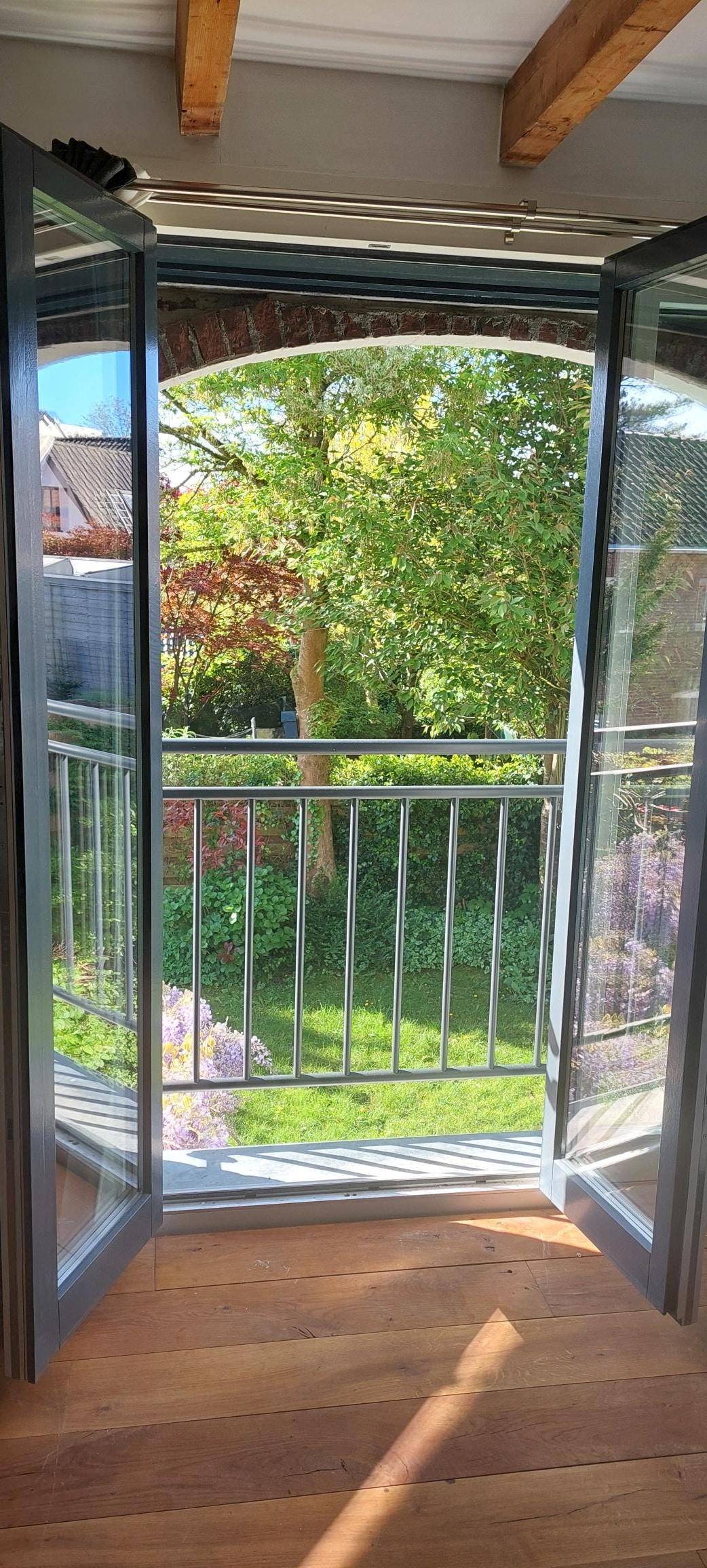 Double doors open to a verdant view of a garden, showcasing lush greenery and colorful plant life. Sunlight gently filters through, enhancing the natural beauty of the outdoor space. A wooden floor is visible inside the room, adding a warm touch to the interior.
