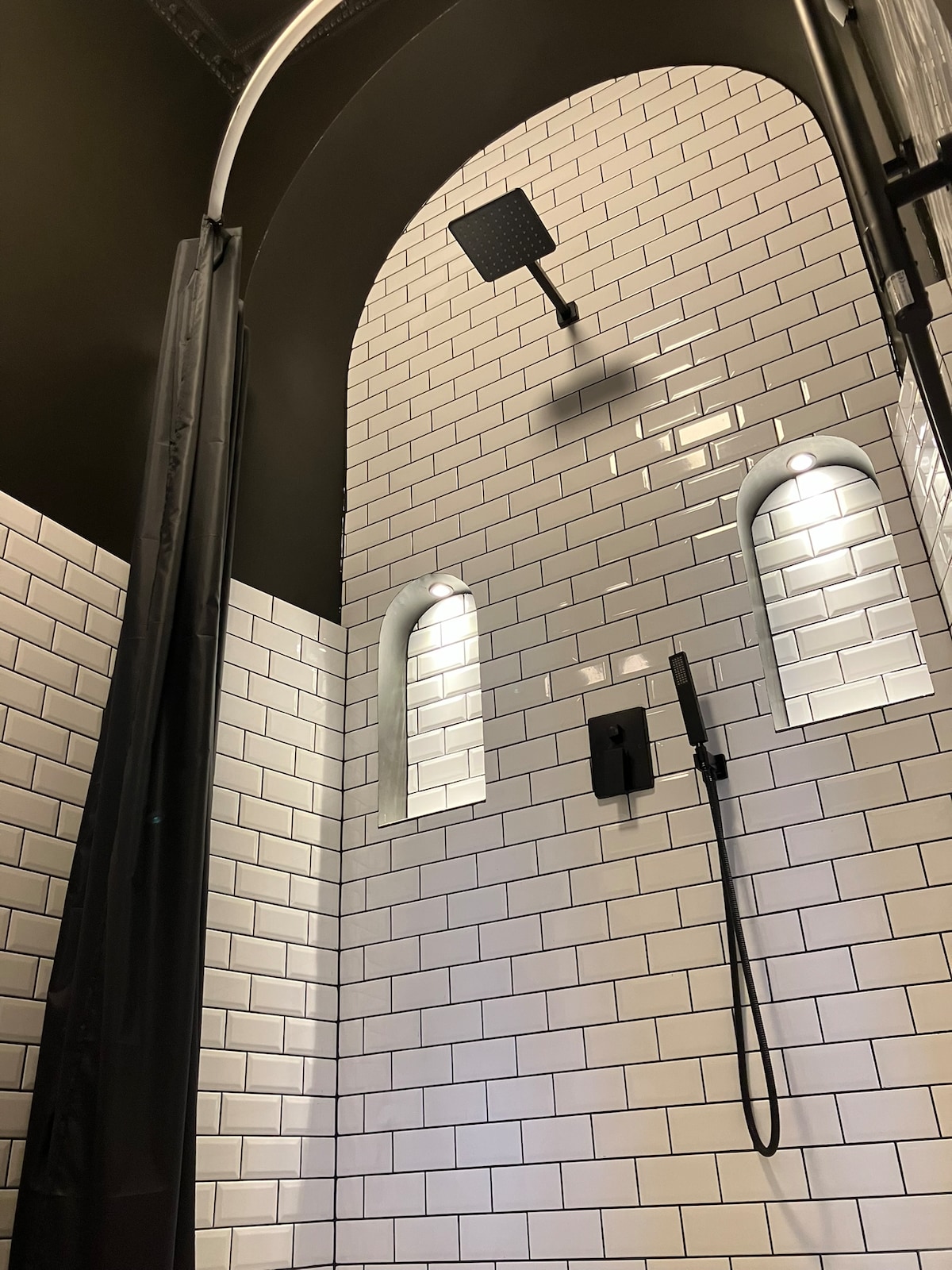 The bathroom features a modern shower area, characterized by stylish white subway tiles and a sleek black ceiling. An overhead rain showerhead is positioned at the center, complemented by two recessed wall niches with soft lighting, creating a contemporary yet understated aesthetic.