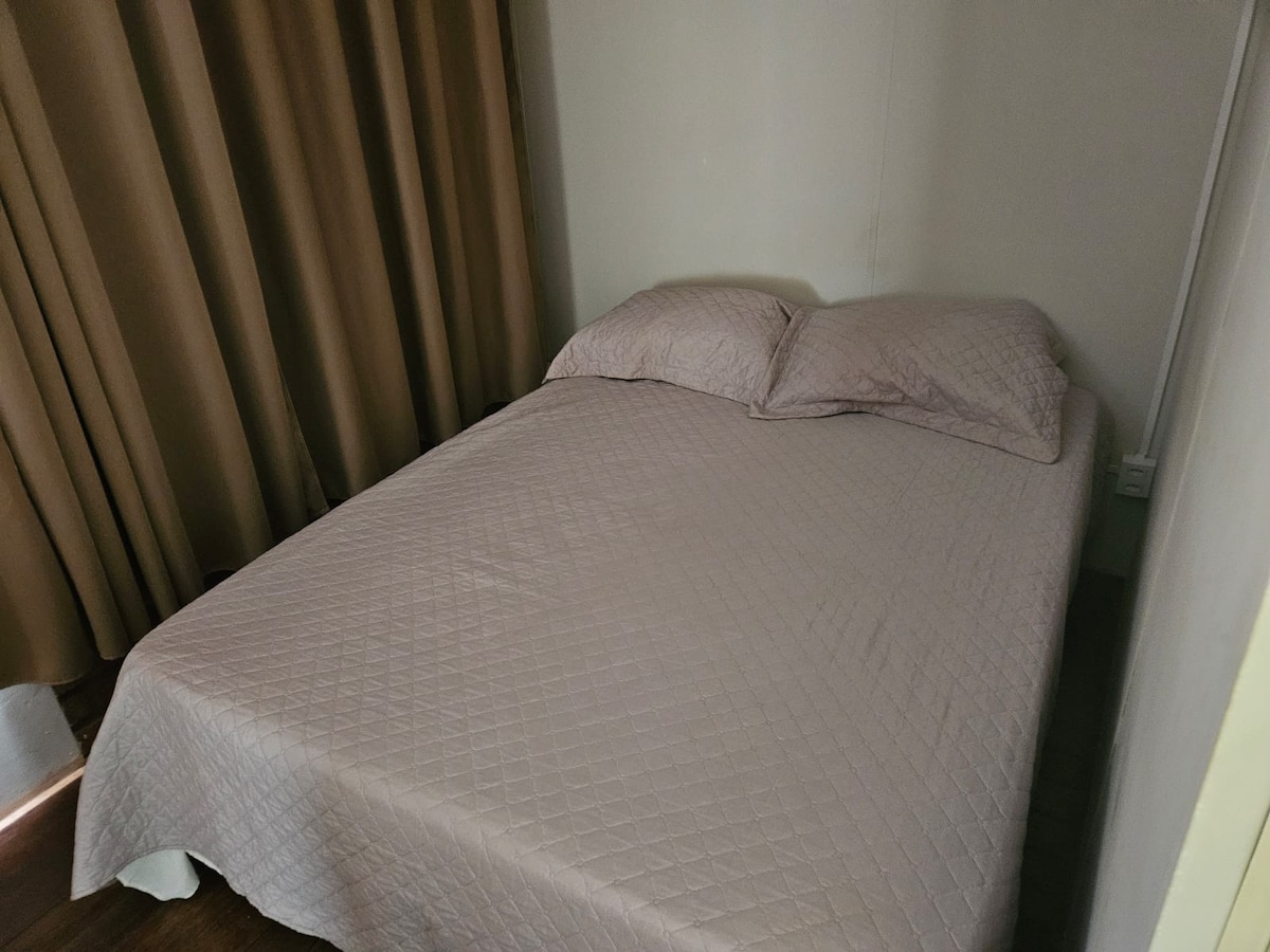 A spacious bed with a soft, quilted blanket is positioned against a wall. Two pillows are neatly arranged at the head of the bed. Light filtering in from a nearby window highlights the smooth, dark wooden floor and the muted curtains providing privacy.