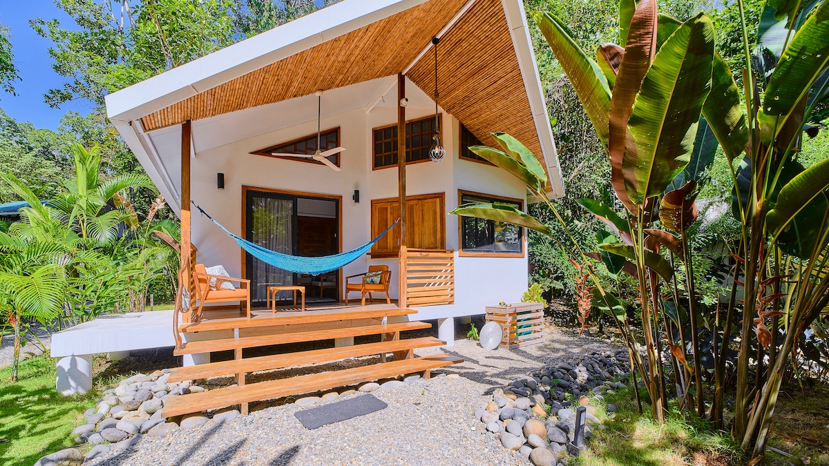 A welcoming exterior is showcased, featuring a covered porch with a hammock and wooden seating. Lush tropical plants frame the entrance, while pebbles create a textured path leading to the door. Natural light filters through large windows, enhancing the serene atmosphere.