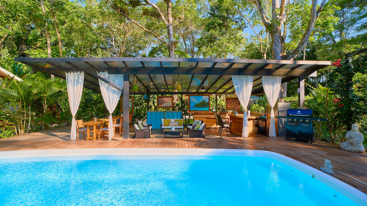 An outdoor dining area is situated beside a tranquil pool, featuring a wooden deck surrounded by lush greenery. The space is covered by a stylish roof and includes a gas grill, comfortable seating, and dining tables, creating a relaxing atmosphere for gatherings.