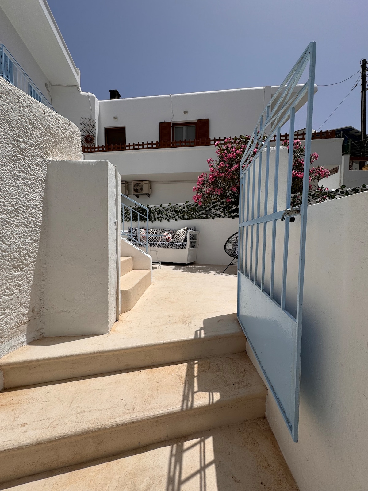 A light blue gate opens to reveal a staircase leading up to a private yard. Blooming flowers frame the entrance, and a cozy seating area with cushions is visible at the top. Bright sunlight enhances the clean, white walls and steps.