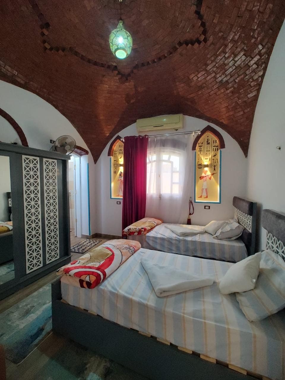 The room features two spacious beds adorned with striped linens and white pillows. A decorative mirror stands against the wall, and vibrant wall art is visible beside the window, allowing natural light to fill the space. A unique arched ceiling is also present.