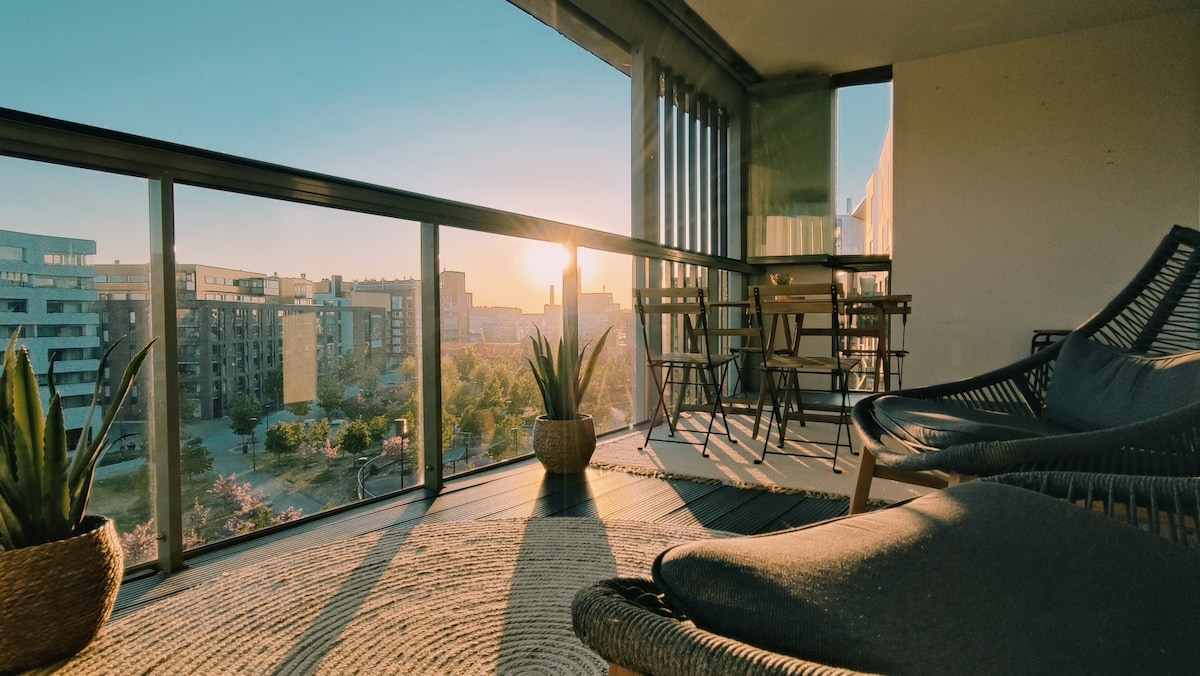A spacious balcony features views of the surrounding park at sunset. Comfortable seating arrangements with stylish chairs and potted plants are present. Sunlight casts a warm glow on the floor, highlighting the inviting atmosphere. The expansive glass doors allow for easy access to this outdoor space.