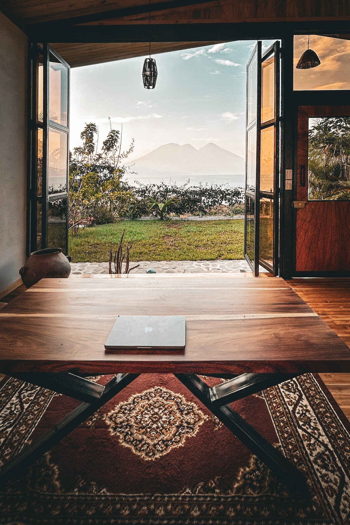 A wooden table is positioned in front of large glass doors, which open to reveal scenic views of the lake and distant mountains. Natural light fills the space, highlighting the rich texture of the hardwood floor and a patterned rug beneath the table.