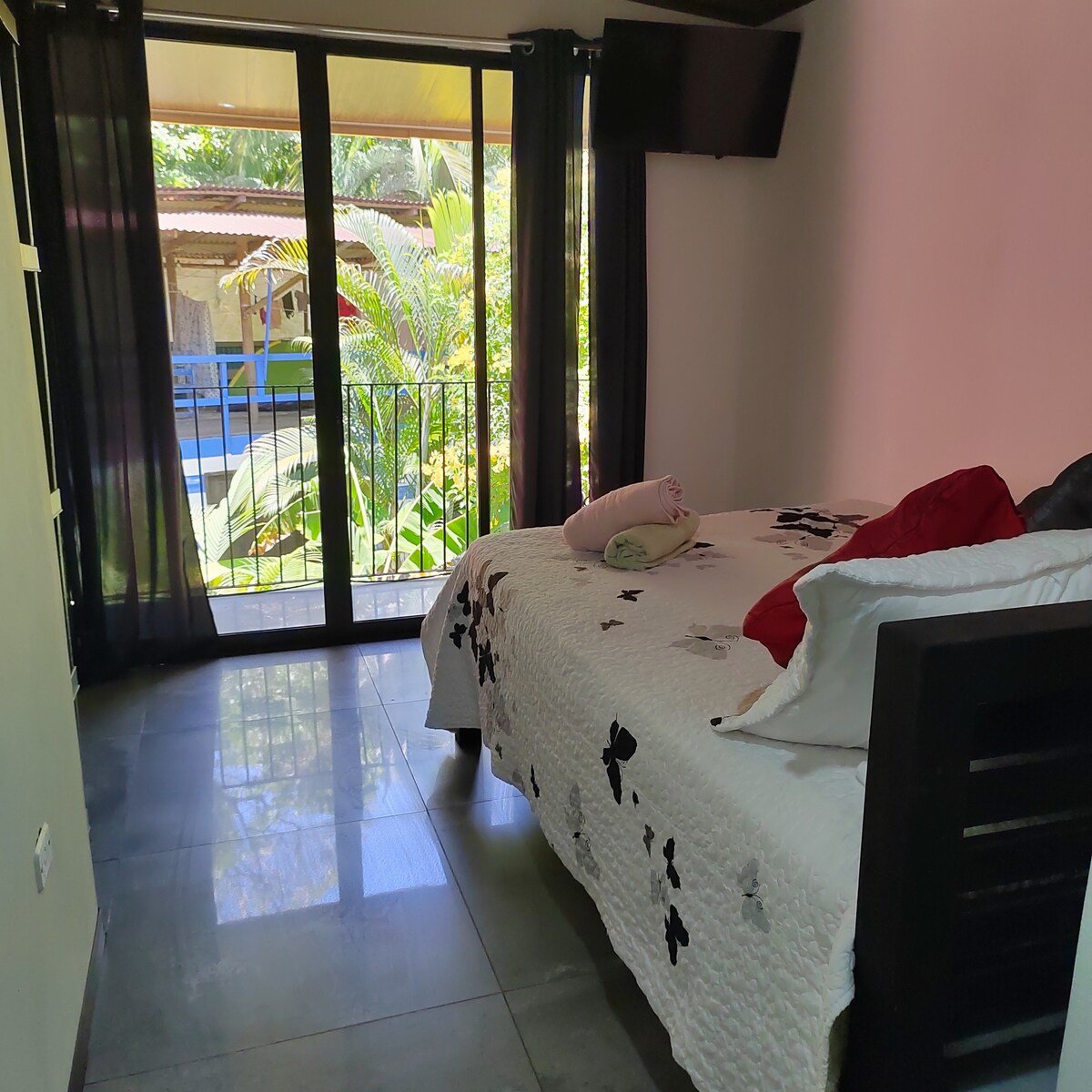 A spacious bedroom is shown featuring a large bed covered with a white quilt decorated with black butterflies. Natural light enters through sliding glass doors leading to a balcony, revealing views of lush greenery and a pool area beyond.