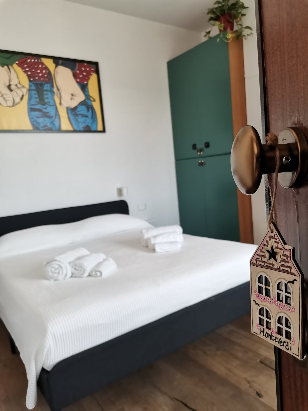 A spacious sleeping area is presented with a neatly made bed, complete with fresh towels arranged on the end. A vibrant piece of wall art featuring footwear adds a colorful touch. A wooden door with a decorative keychain hangs nearby.