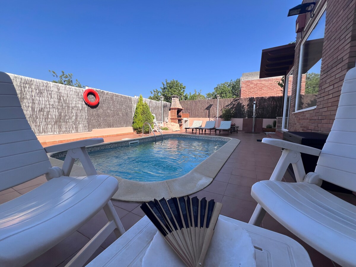 A private pool area is visible, featuring a rectangular swimming pool surrounded by tiled flooring. Two white lounge chairs are positioned near the water, complemented by a small table. A tree and a brick structure can be seen in the background, creating a tranquil setting.
