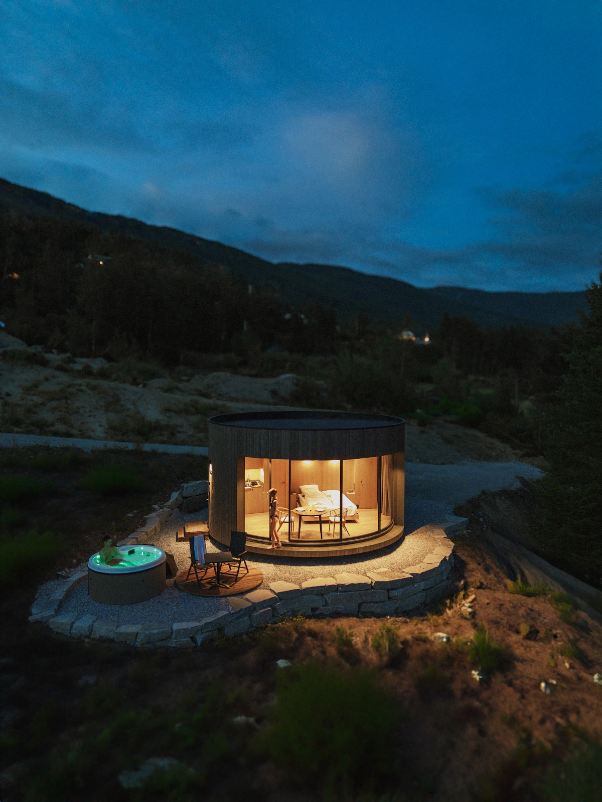 A modern cabin is illuminated in warm light against a twilight backdrop. The circular structure features large windows offering views of surrounding nature. A private jacuzzi is situated nearby, adding to the serene outdoor space designed for relaxation.