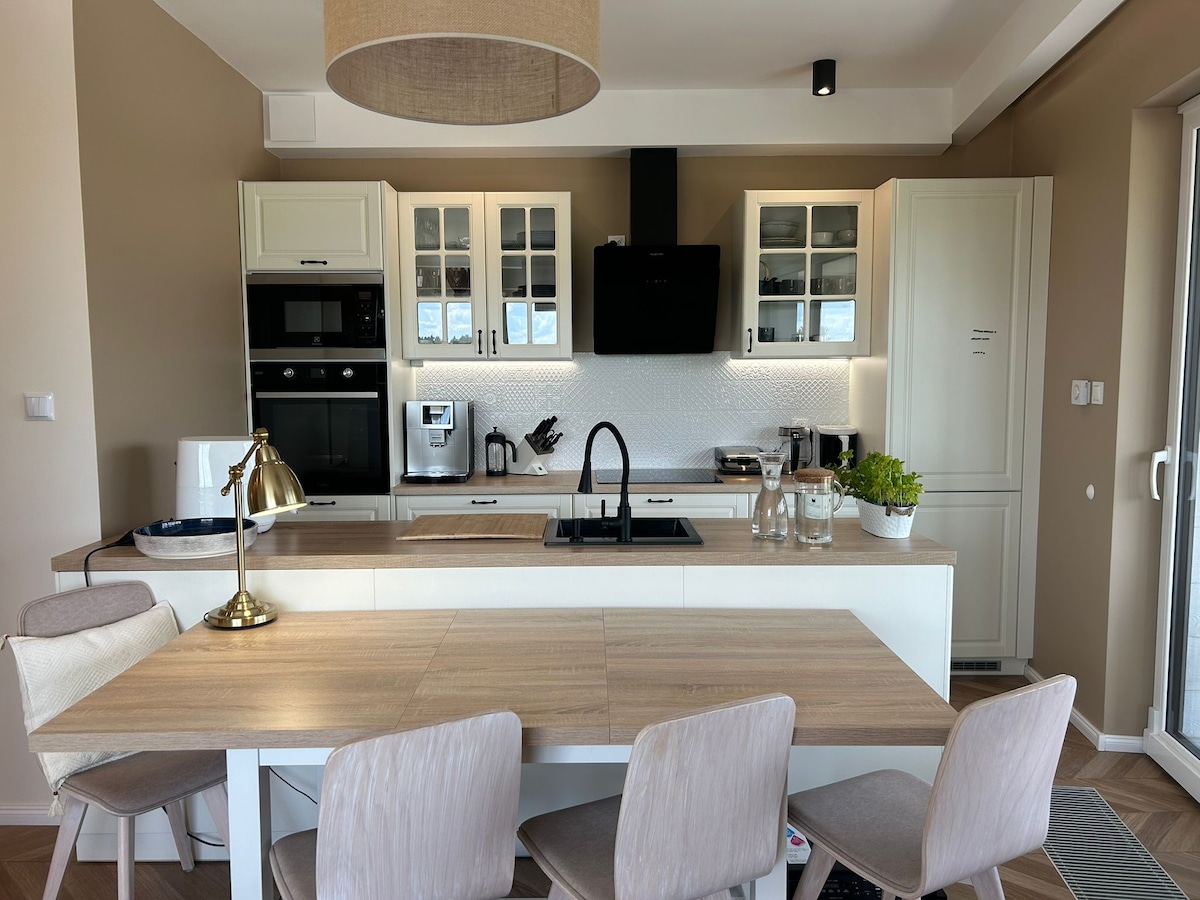 A spacious kitchen features modern appliances, ample cabinetry, and a sleek countertop. A dining table with light-colored chairs is positioned near a window, providing a bright atmosphere. A subtle decorative plant and a stylish lamp add a touch of warmth to the space.