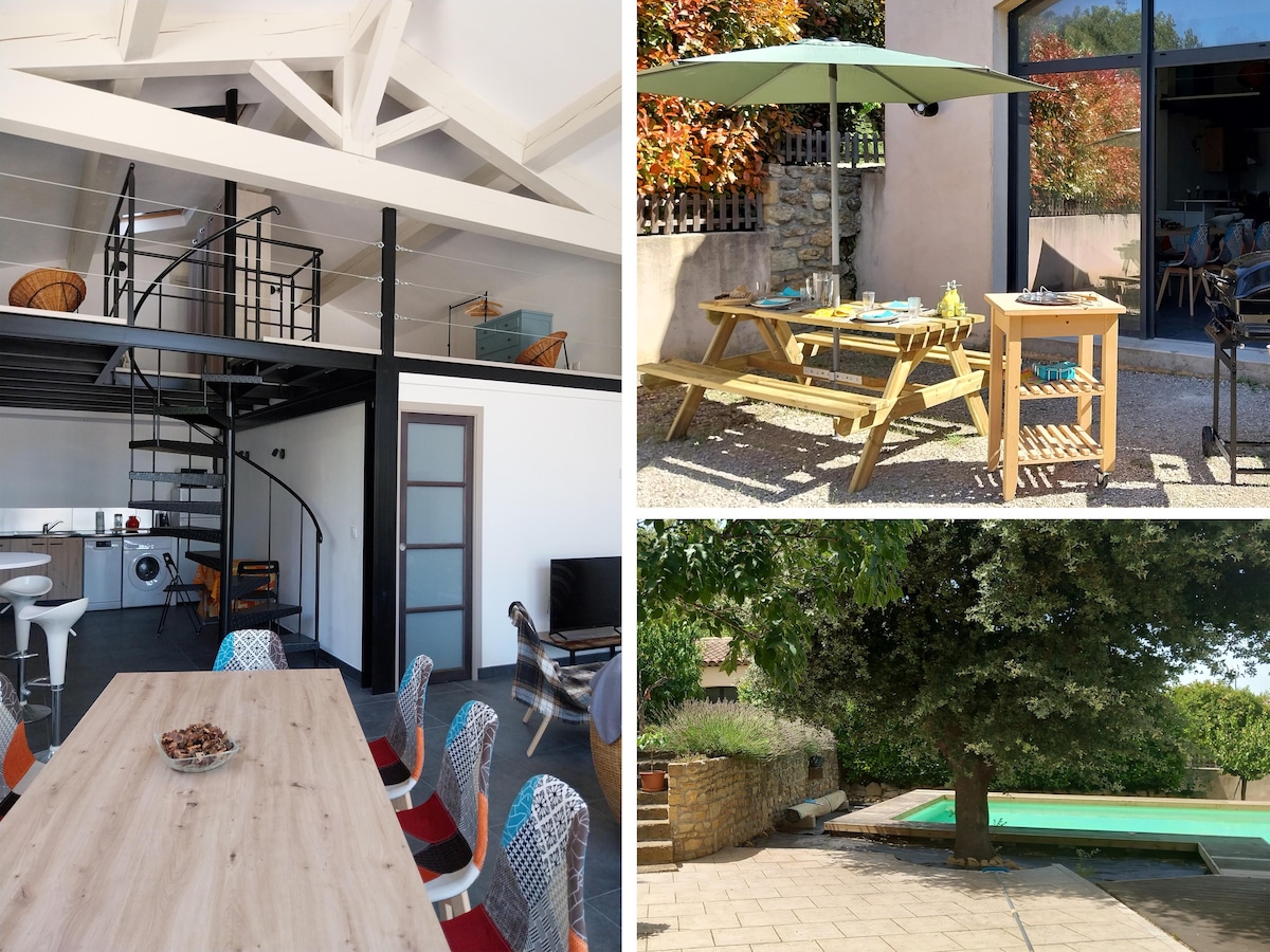The loft's interior features a spacious open-plan living area with a modern kitchen and dining table. A spiral staircase leads to the mezzanine bedrooms. The outdoor space includes a picnic-style dining table under an umbrella, with a view of the shared pool surrounded by greenery.