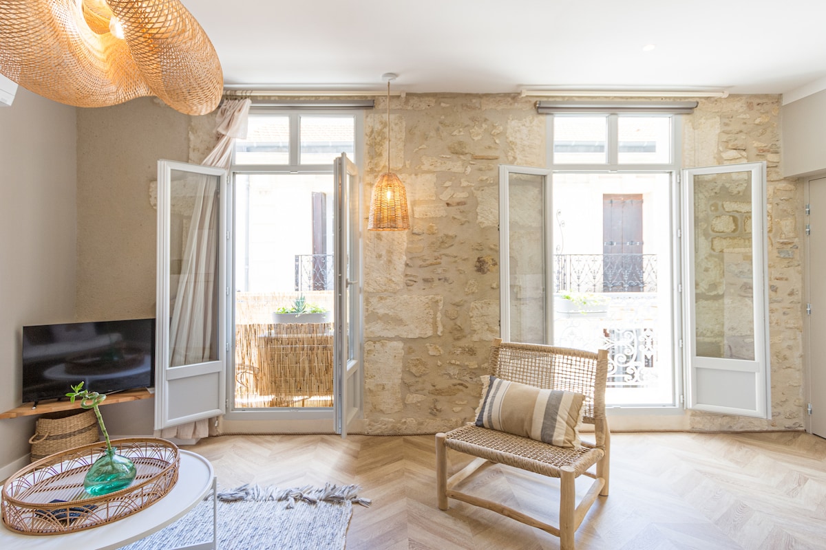 The spacious living area features natural light flowing through open windows, highlighting the stone walls and warm wood accents. A simple wooden chair is positioned beside a decorative round table, while a small television is mounted on the wall, contributing to a cozy atmosphere.