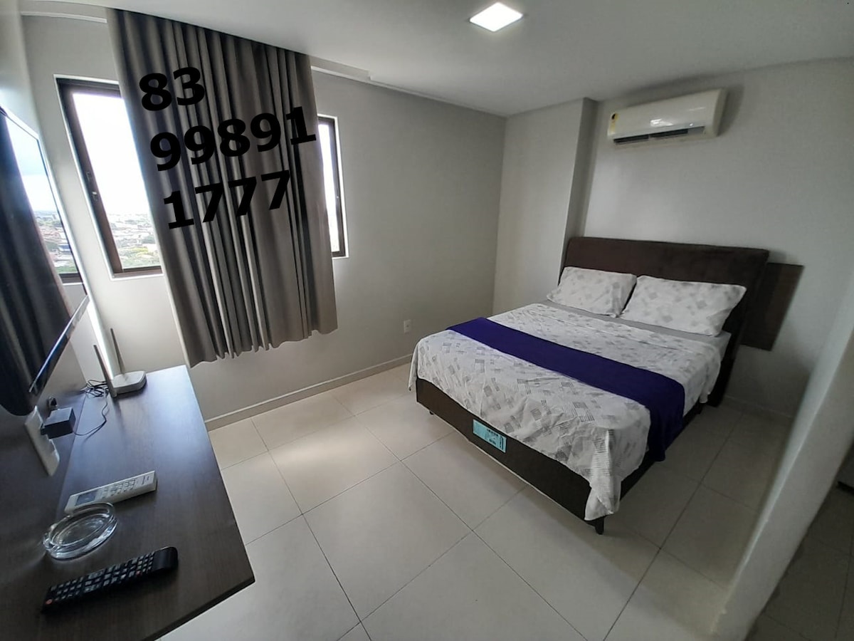 A well-lit bedroom features a double bed with a patterned white and grey duvet. Two windows allow natural light to enter, complemented by neutral-colored curtains. An air conditioning unit is mounted above, and a television is positioned on a low stand nearby.