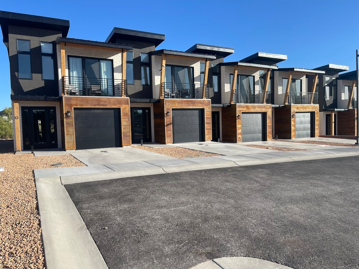 A row of modern townhomes with a sleek design features large windows and a mix of wood and metal exteriors. Each unit is complemented by a spacious driveway, while a clear blue sky enhances the fresh appearance of the building structure.