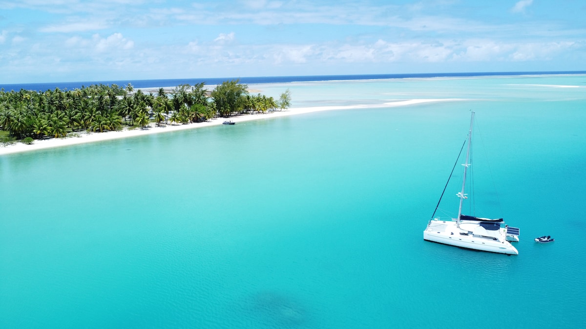 A catamaran is anchored in a turquoise lagoon, surrounded by lush greenery and a sandy shore. The calm water reflects the sky, enhancing the serene atmosphere of this tropical setting. An additional small boat is visible near the shoreline.