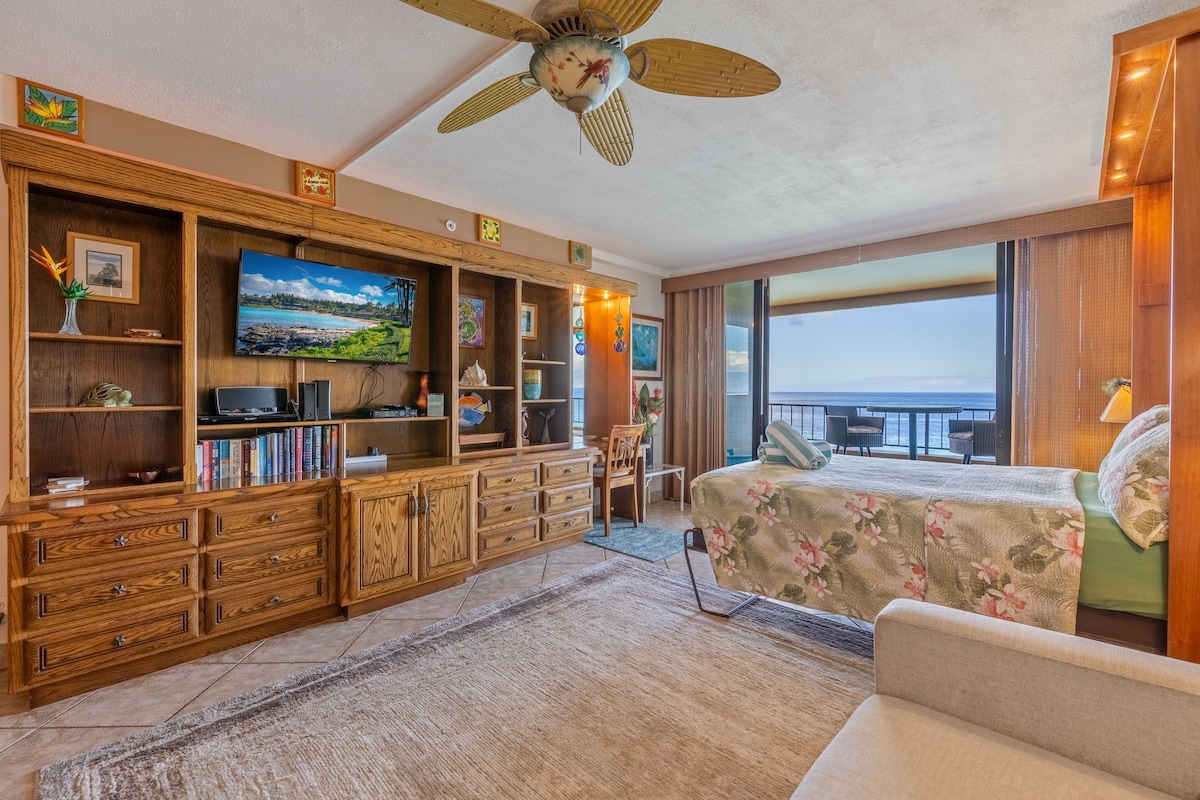 A spacious studio interior showcases wooden cabinetry and a flat-screen TV. Natural light filters through large sliding glass doors, leading to a private lanai with ocean views. The sleeping area features a queen-size bed adorned with floral bedding, complemented by a cozy sitting area with a light-colored couch.