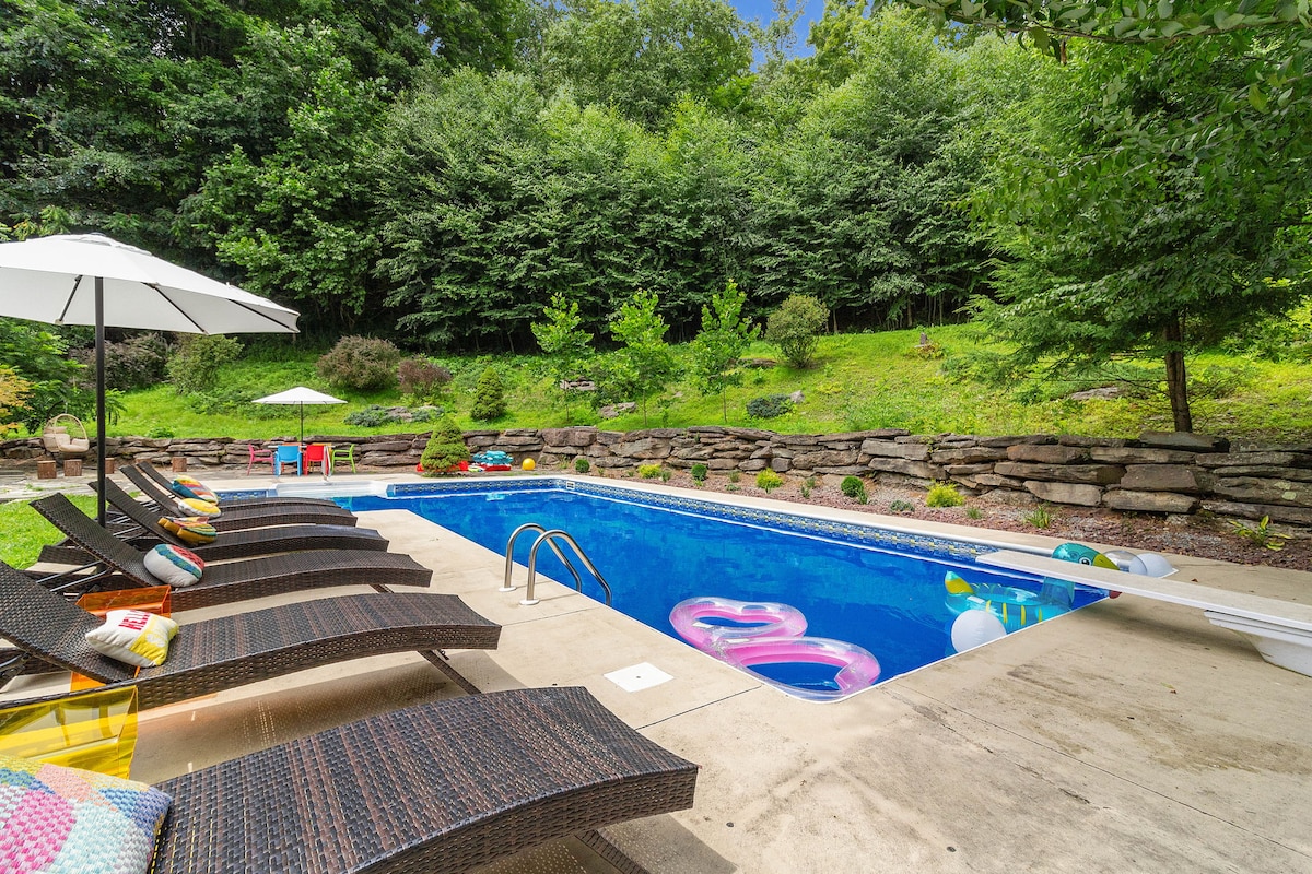 An inground, L-shaped heated pool is surrounded by a spacious concrete deck, featuring comfortable lounge chairs and colorful cushions. Shade is provided by an umbrella, and greenery surrounds the area, creating a serene outdoor environment. Inflatable pool toys can be seen floating in the water.