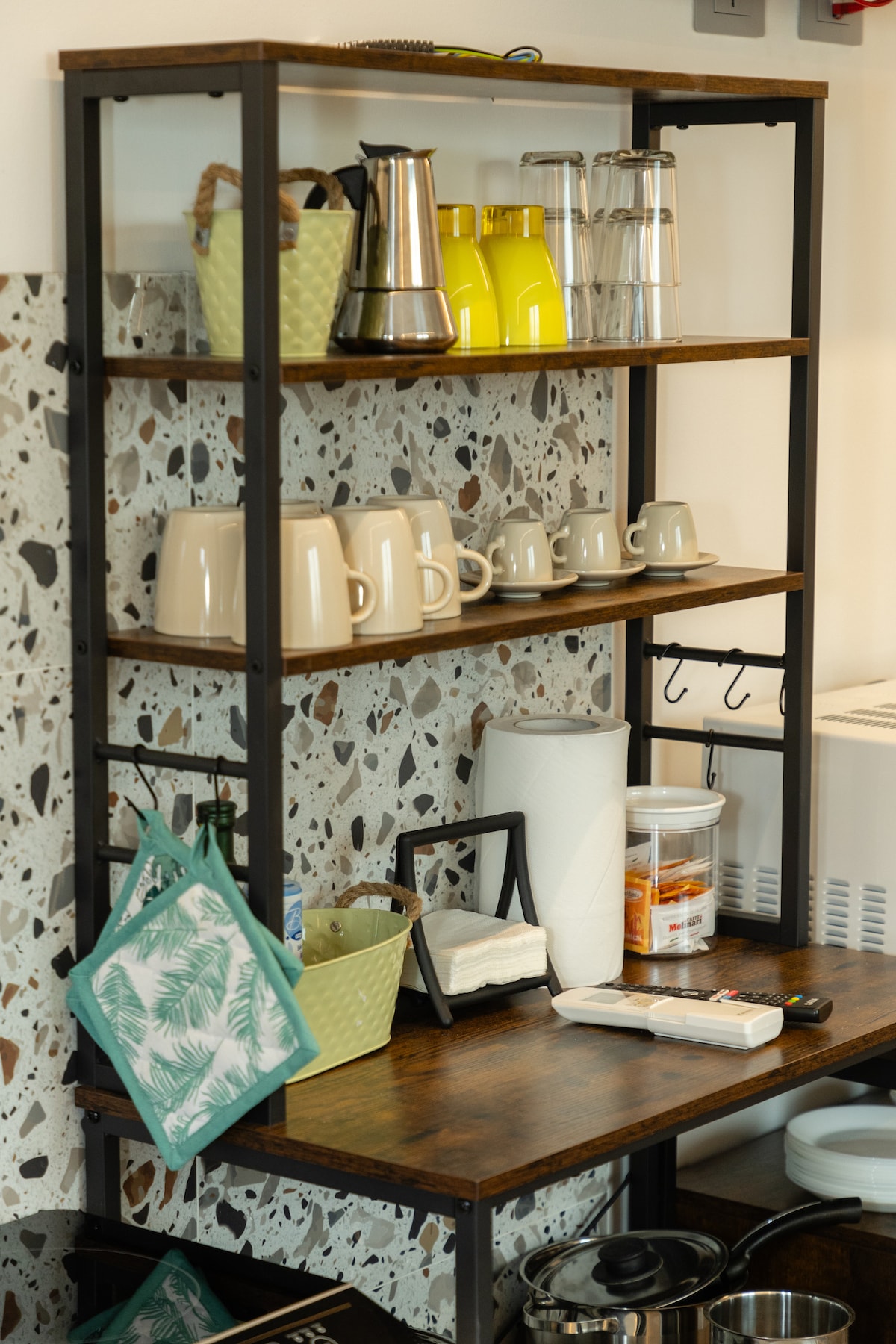 A well-organized kitchen corner is displayed on a multi-level shelving unit made of wood and metal. Various glassware, stylish mugs, and kitchen supplies are neatly arranged, along with two colorful baskets. A decorative towel hangs from the rack, contributing to the functional space.