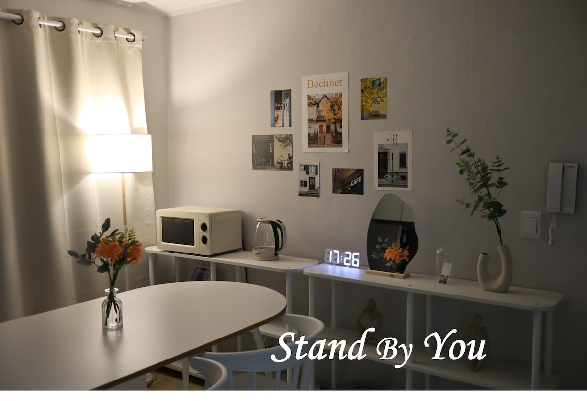 A bright dining area is showcased with a simple white table and chairs. The wall features a collage of photographs. A microwave and kettle are positioned on a shelf, with a clock and mirror nearby. Soft lighting is provided by a floor lamp in the corner.