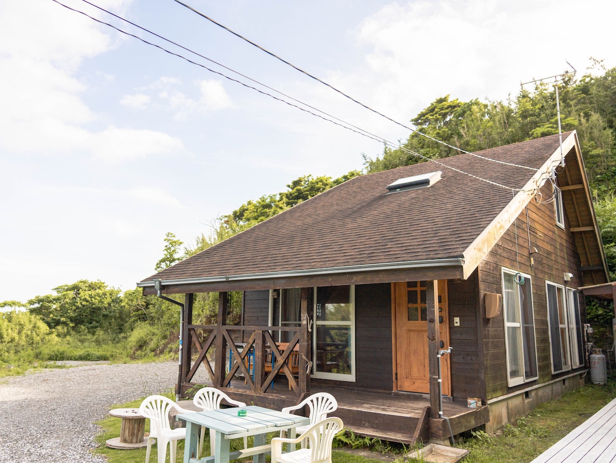 A two-story log house features a wooden exterior and a covered porch area. Outdoor seating is provided with white chairs around a table. Surrounding greenery is visible, and the clear sky adds to the tranquil setting of the house.