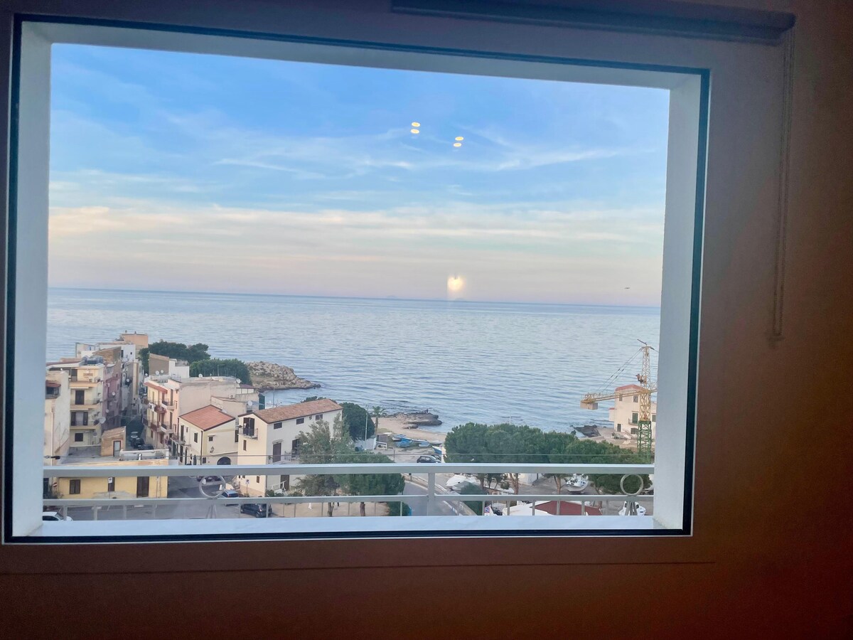 A large window frames a view of the tranquil sea, where the sun sets over the horizon. The coastline features small buildings and a harbor, highlighted by soft colors of the evening sky, creating a serene and open atmosphere.