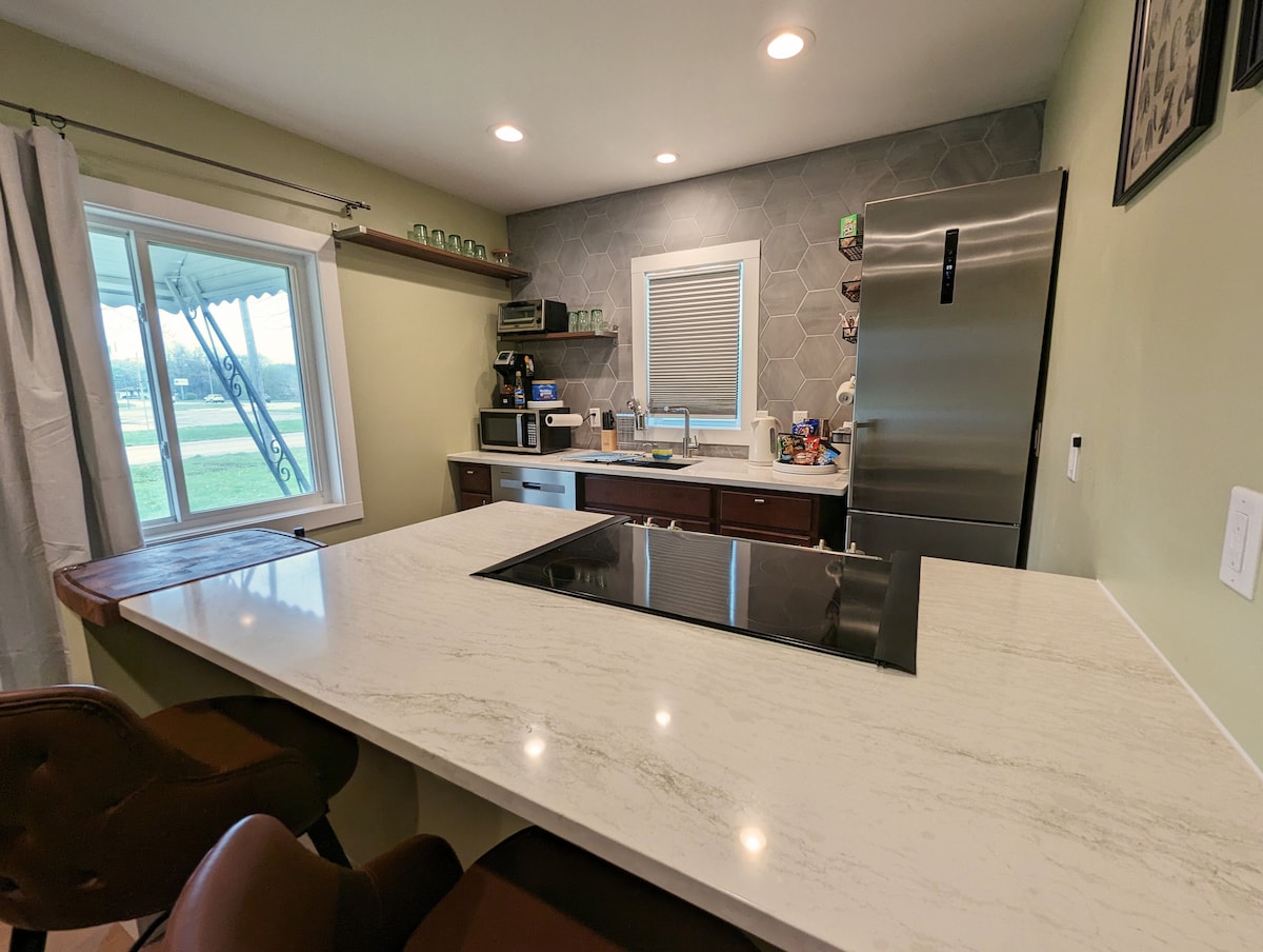 A modern kitchen features a quartz countertop with a built-in stovetop, complemented by dark cabinetry. Stainless steel appliances, including a refrigerator and microwave, are present. Natural light comes through a window overlooking the exterior, while comfortable seating is available at the island.